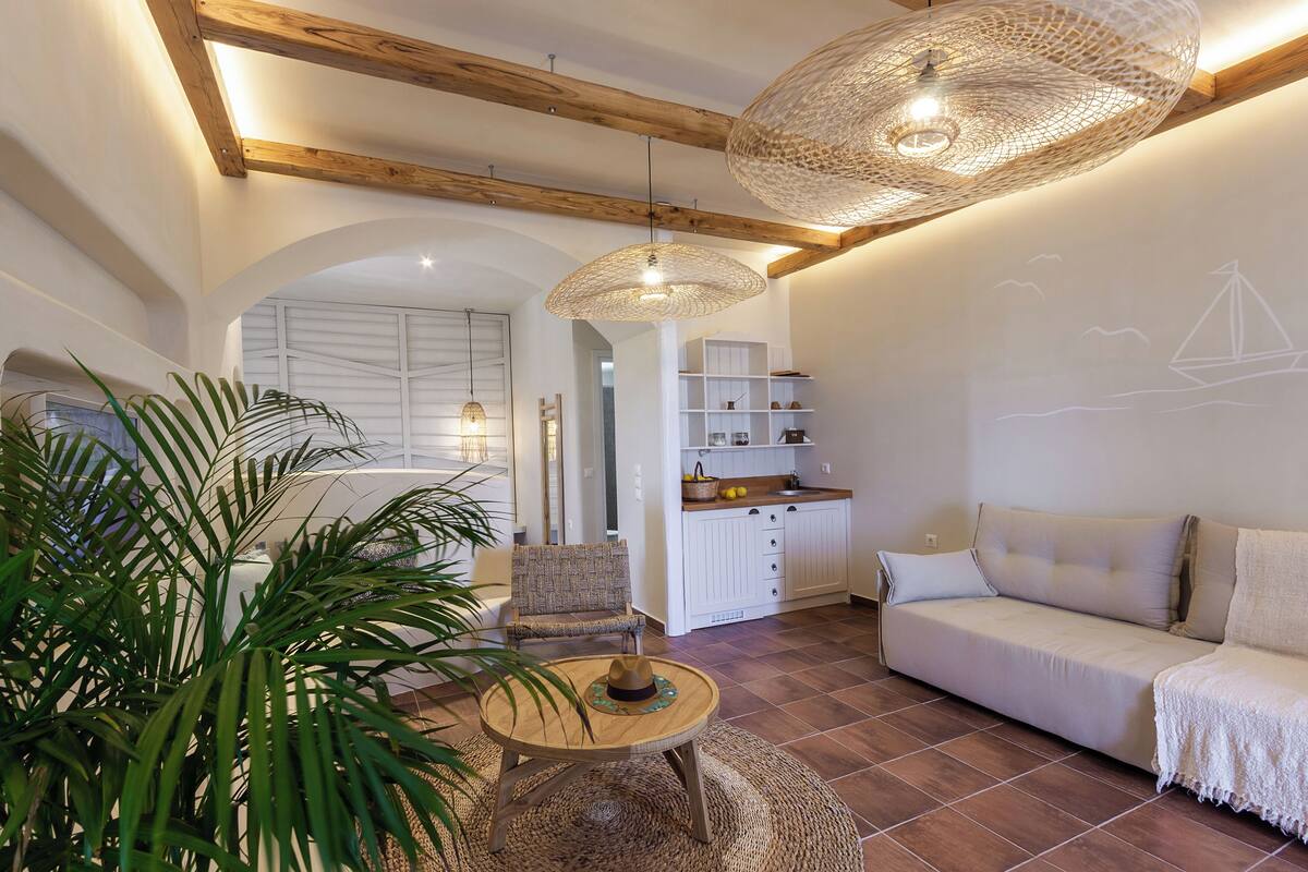 A living room with a sofa, armchair, and coffee table, a kitchen in the background, and a large plant.