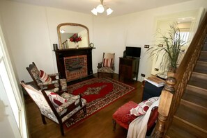 A living room with a fireplace, patterned chairs, a red rug, and a wooden staircase.