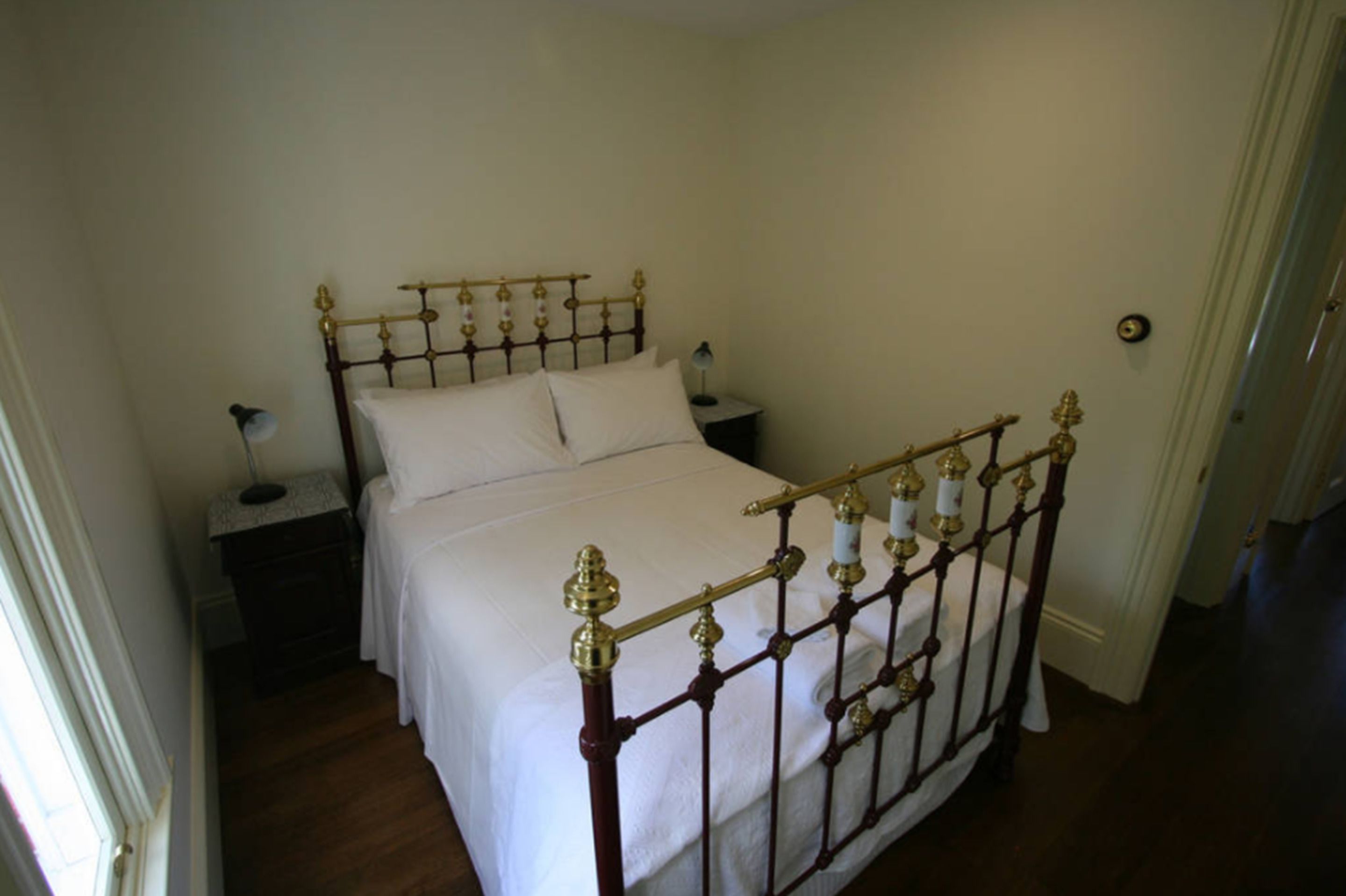 A single bed with a wooden frame and brass accents, a nightstand with a lamp, and a plain wall.