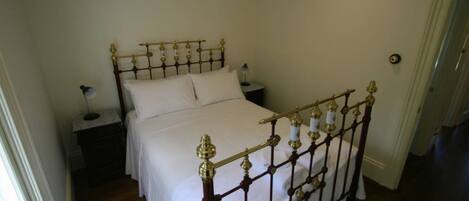 A single bed with a wooden frame and brass accents, a nightstand with a lamp, and a plain wall.