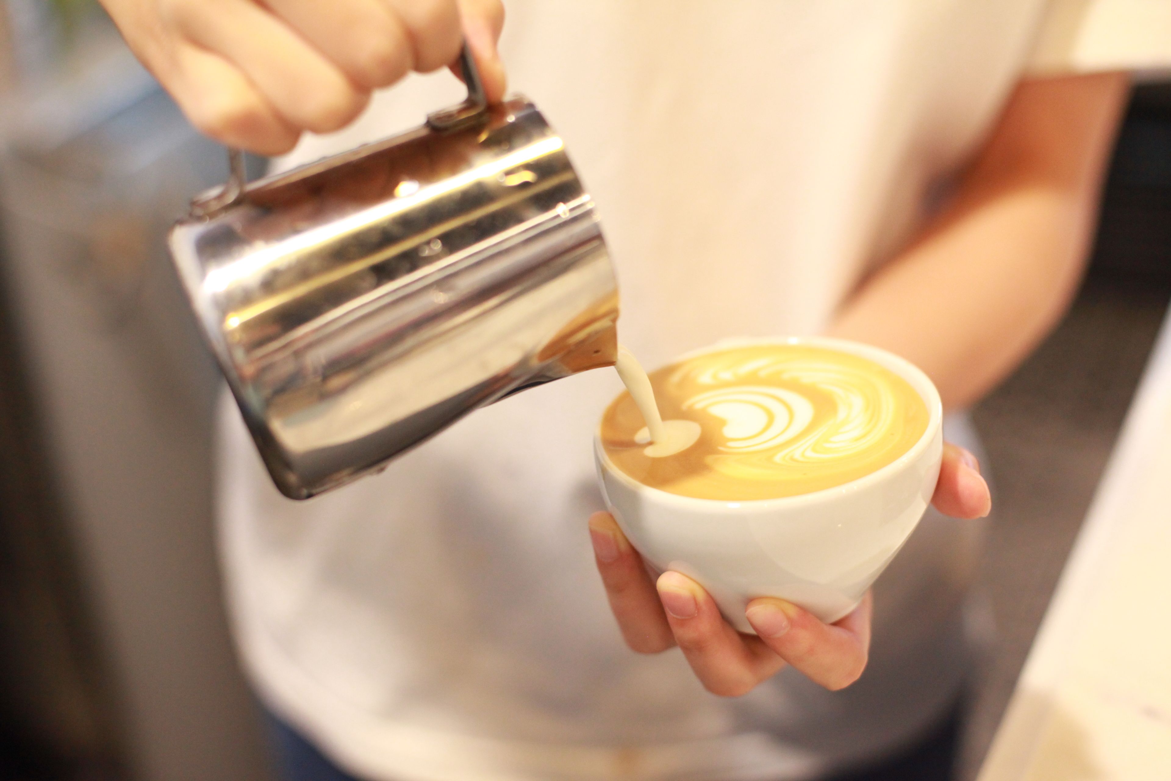 A person pouring milk into a cup of coffee, creating a latte art pattern.
