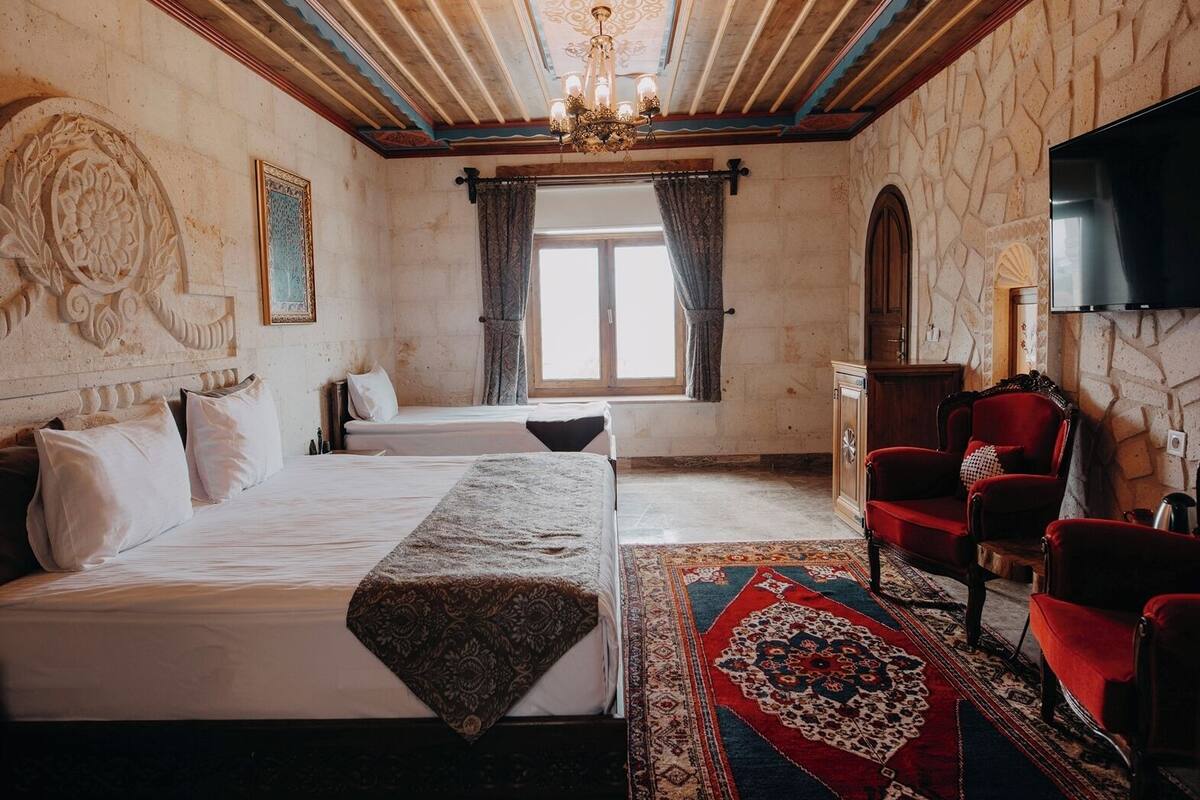 Une chambre avec deux lits, un grand lustre décoratif, un mur en pierre orné de motifs sculptés, un fauteuil rouge et un téléviseur à écran plat fixé au mur.