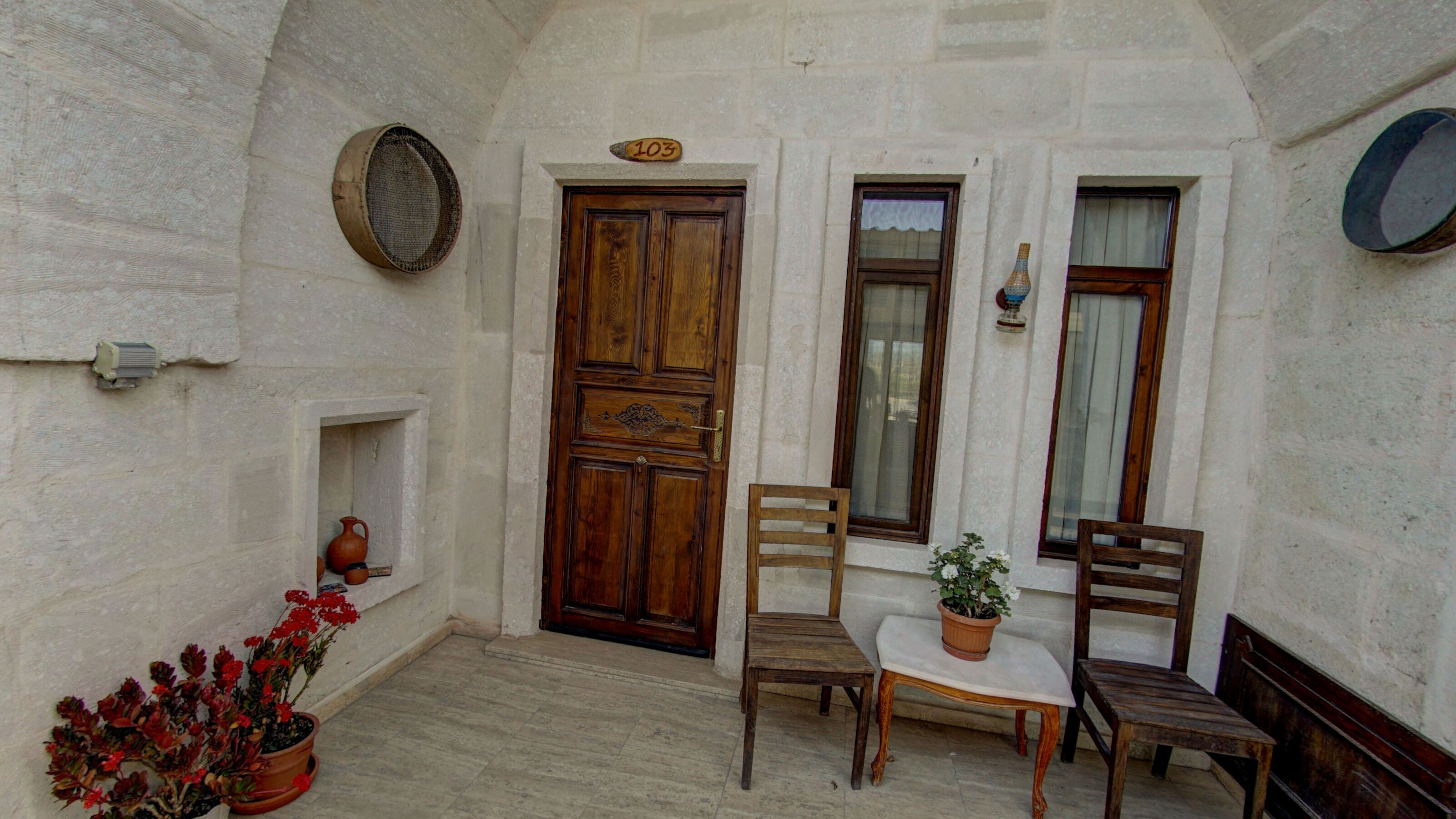 A small courtyard with a wooden door, two windows, a bench, a table, and a potted plant.