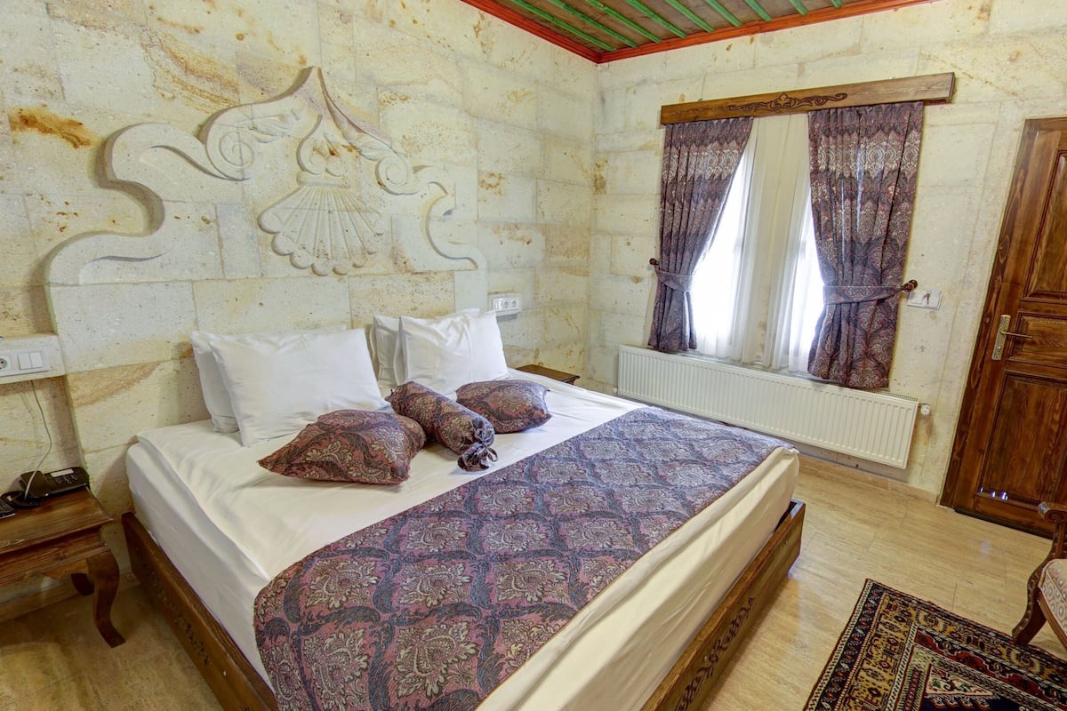 Une chambre à coucher comprenant un lit, une table de chevet, une armoire en bois, une fenêtre avec des rideaux et un tapis à motifs.