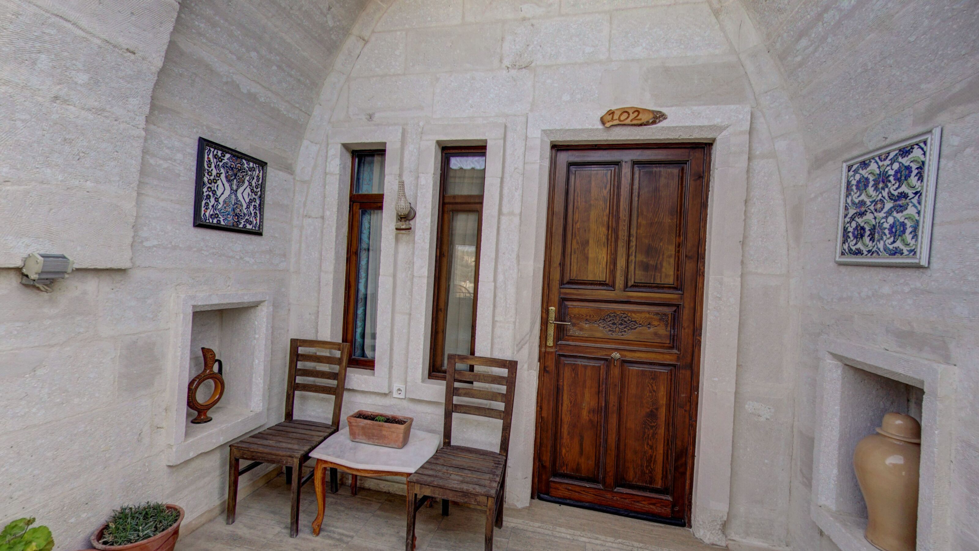 A narrow courtyard with a wooden door marked "102", two wooden chairs, a small table, and a potted plant.