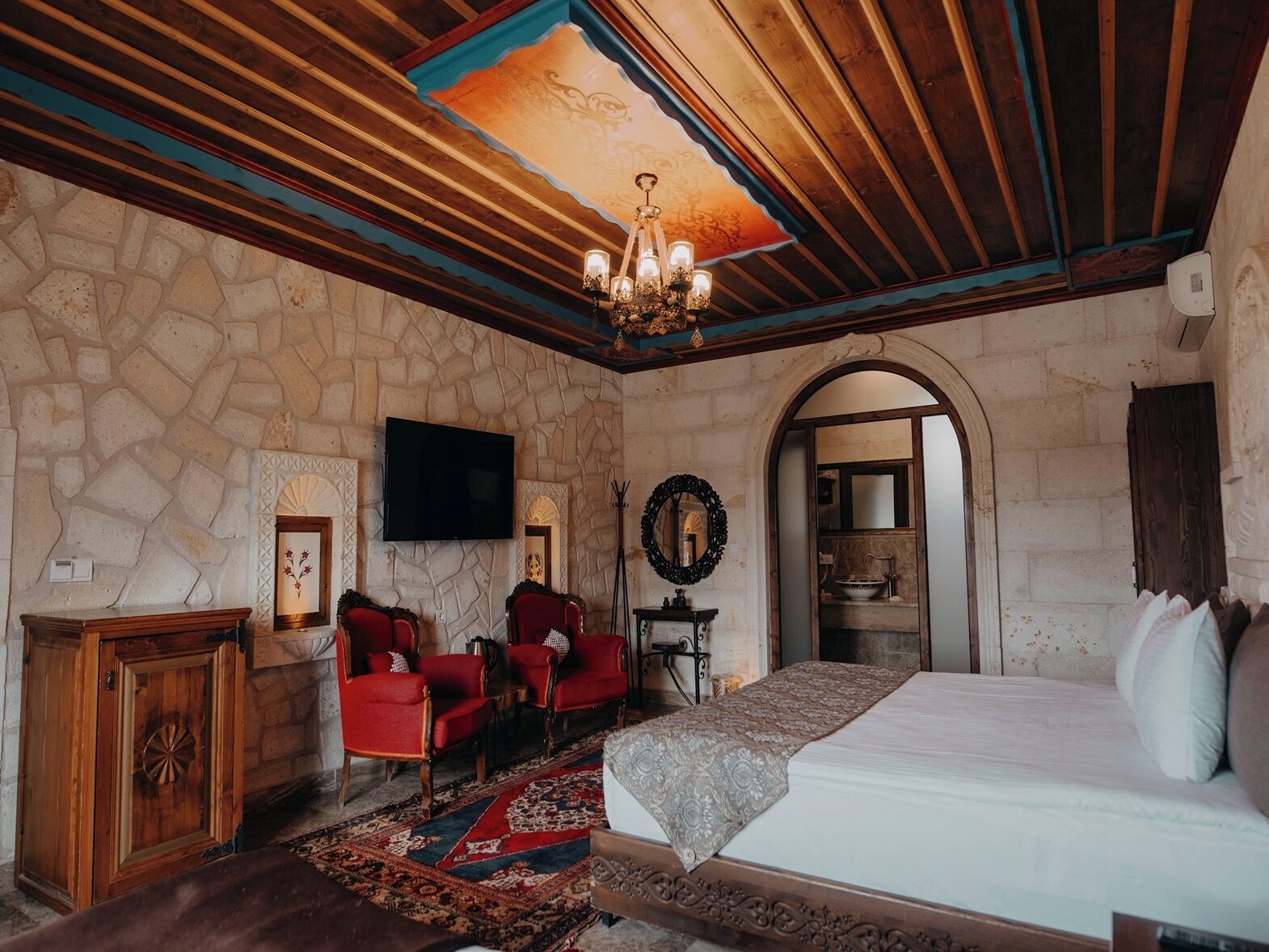 A bedroom with a stone wall, wooden ceiling, a chandelier, a bed, a TV, a mirror, and a red chair.