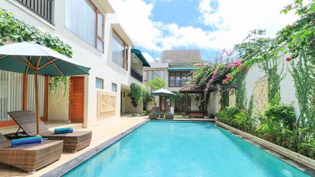A swimming pool with lounge chairs and umbrellas, surrounded by greenery and a building with large windows.