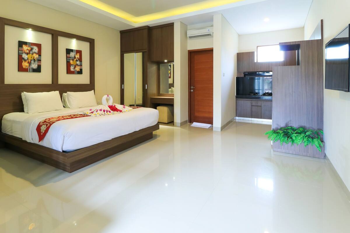 A modern hotel room with a large bed, a wooden wardrobe, a small desk, and a kitchenette.