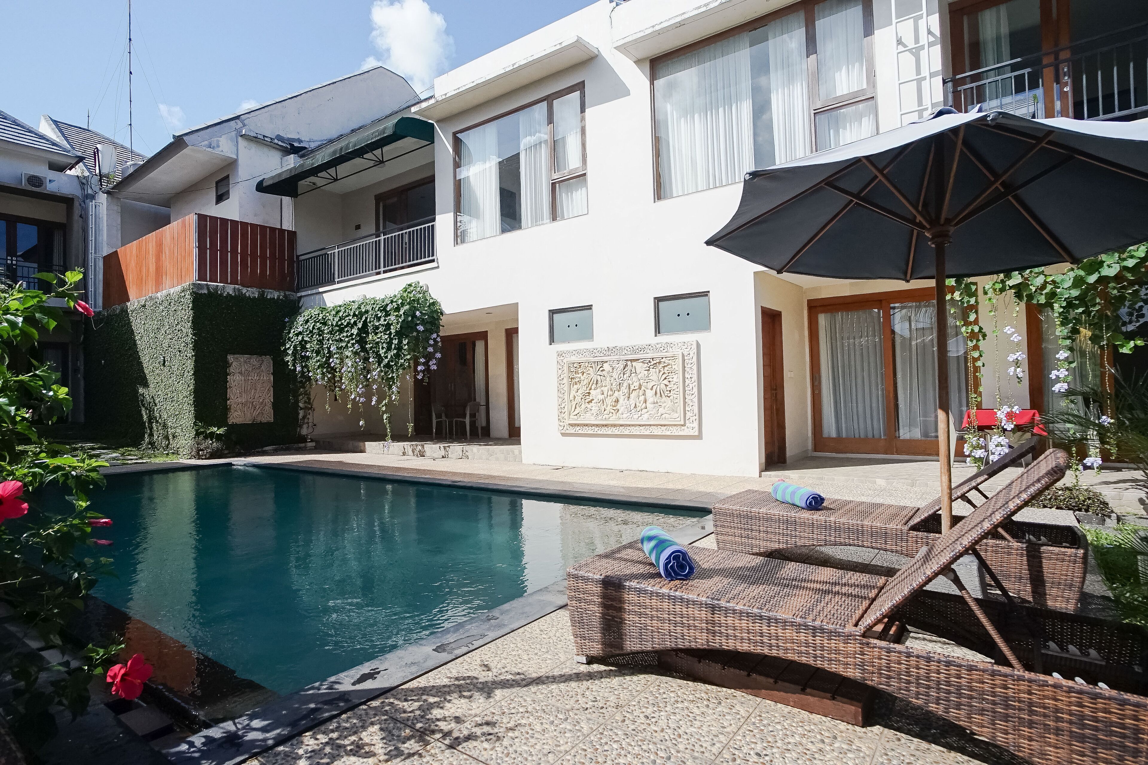 A swimming pool with lounge chairs and an umbrella, surrounded by a white building with balconies and a decorative wall.