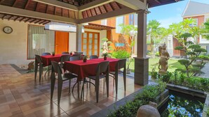 A covered outdoor dining area with tables and chairs, a clock on the wall, and a garden with plants and a statue.