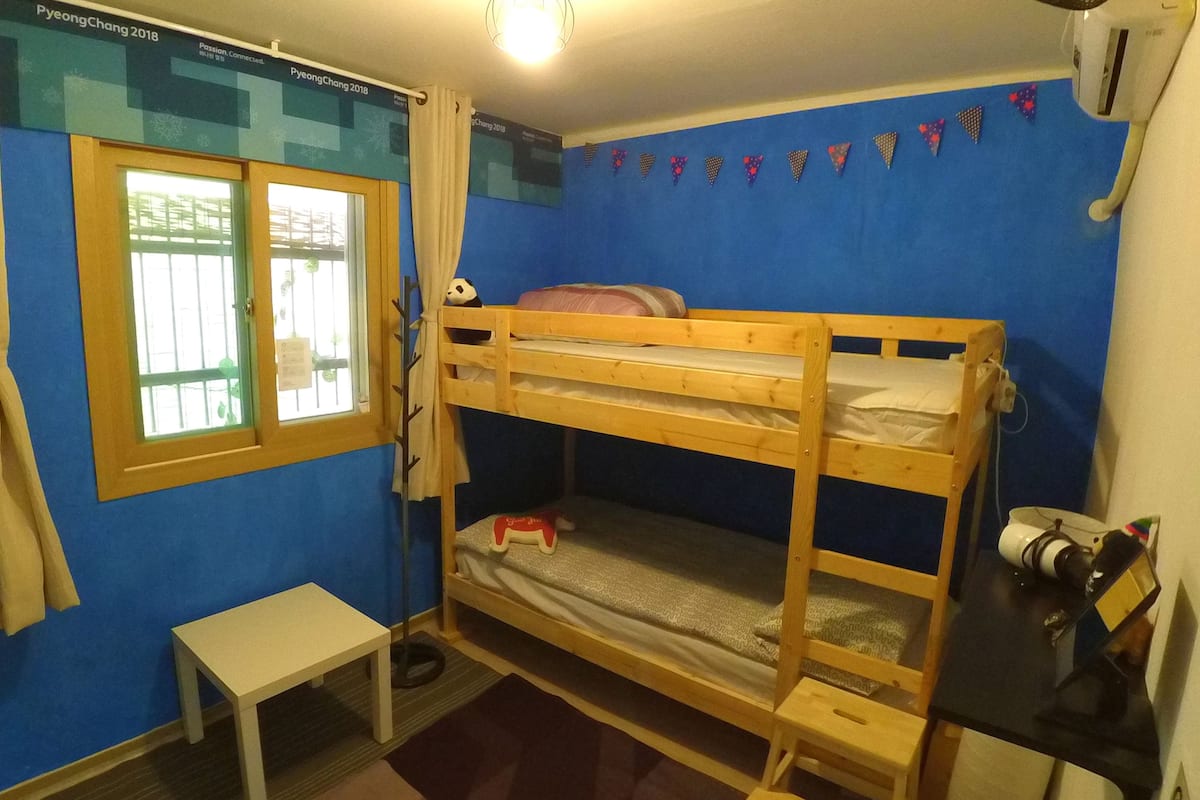 Habitación con literas, una pequeña mesa, una ventana con cortinas y una pared azul con adornos triangulares.
