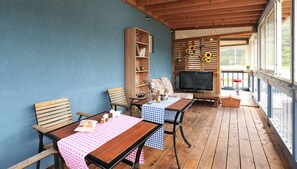 A covered outdoor seating area with wooden tables and chairs, a bookshelf, and a television.