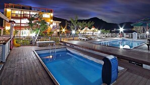Un espace extérieur bien éclairé avec une piscine, une terrasse en bois, des chaises longues et un bâtiment doté de plusieurs balcons.