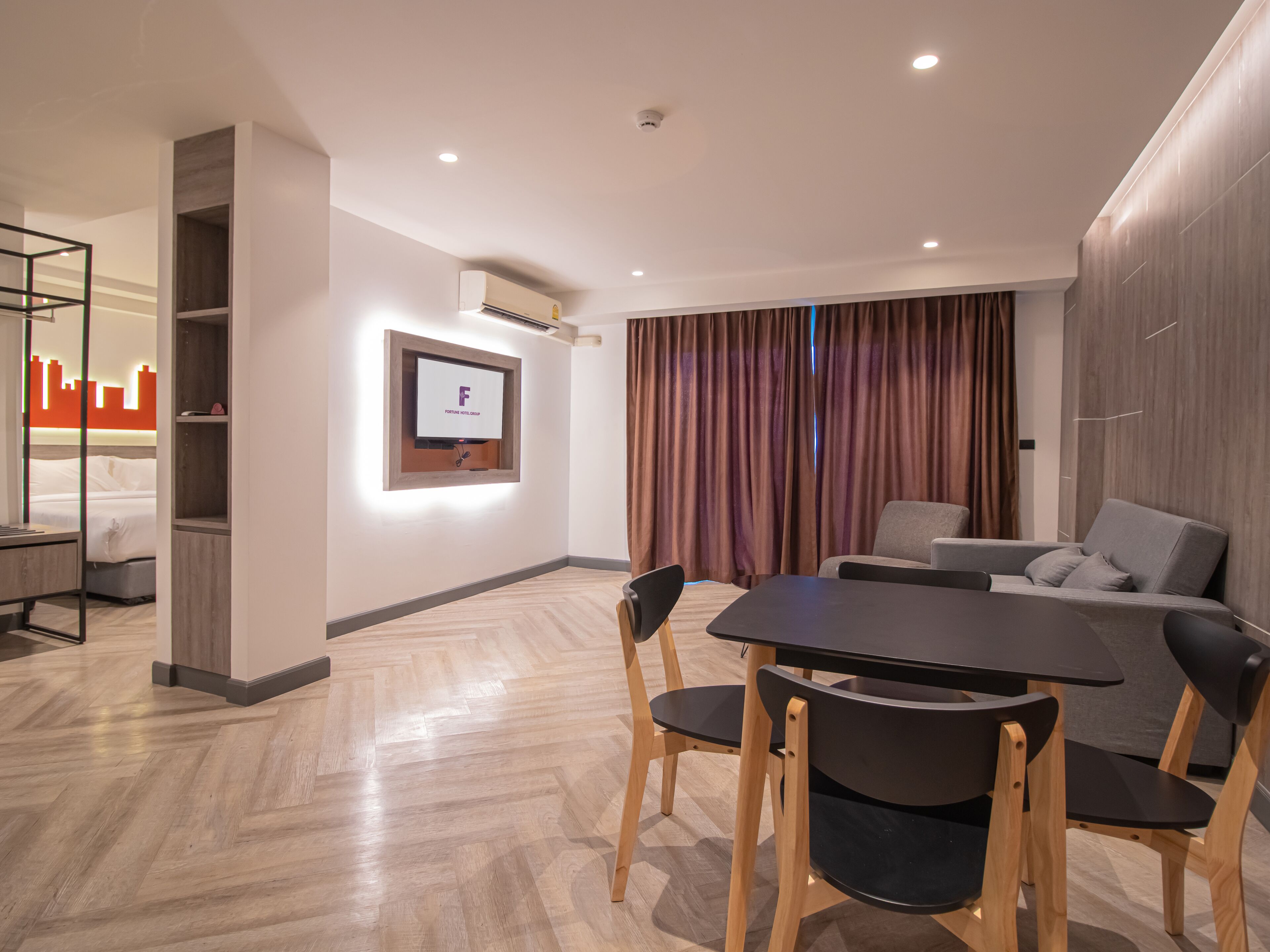 A modern hotel room with a dining area, a flat-screen TV mounted on the wall, and a large window with curtains.