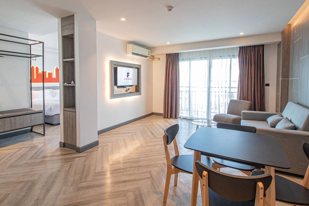 A modern hotel room with a bed, a TV mounted on the wall, a dining area with a table and chairs, and a balcony with curtains.