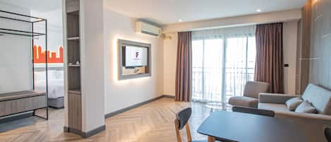 Una habitación de hotel moderna con una cama, un televisor montado en la pared, una zona de comedor con mesa y sillas, y un balcón con cortinas.