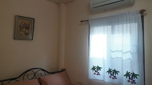 A bedroom with a bed, a wall-mounted air conditioner, a framed picture, and a window with a curtain featuring palm trees and fruit. - Benya Guest House Phimai (Phimai)