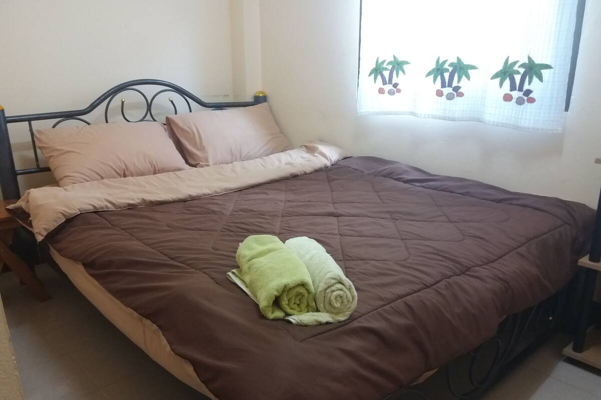 A bed with a brown quilt, a green towel, and a window with a curtain featuring palm trees.
