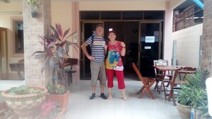 A man and a woman standing in front of a building with a sign that reads "ROOM CHECK IN". There are potted plants and a wooden table with chairs outside. - Benya Guest House Phimai (Phimai)