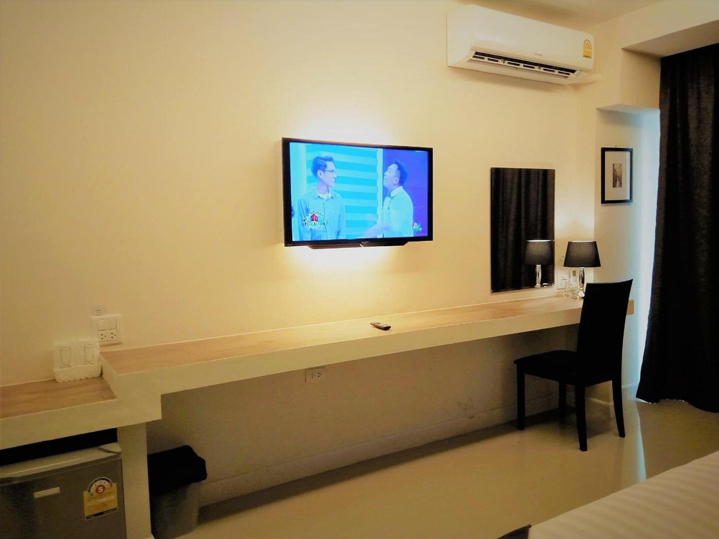 A hotel room with a TV mounted on the wall, a desk with a chair, a small trash bin, and an air conditioning unit.