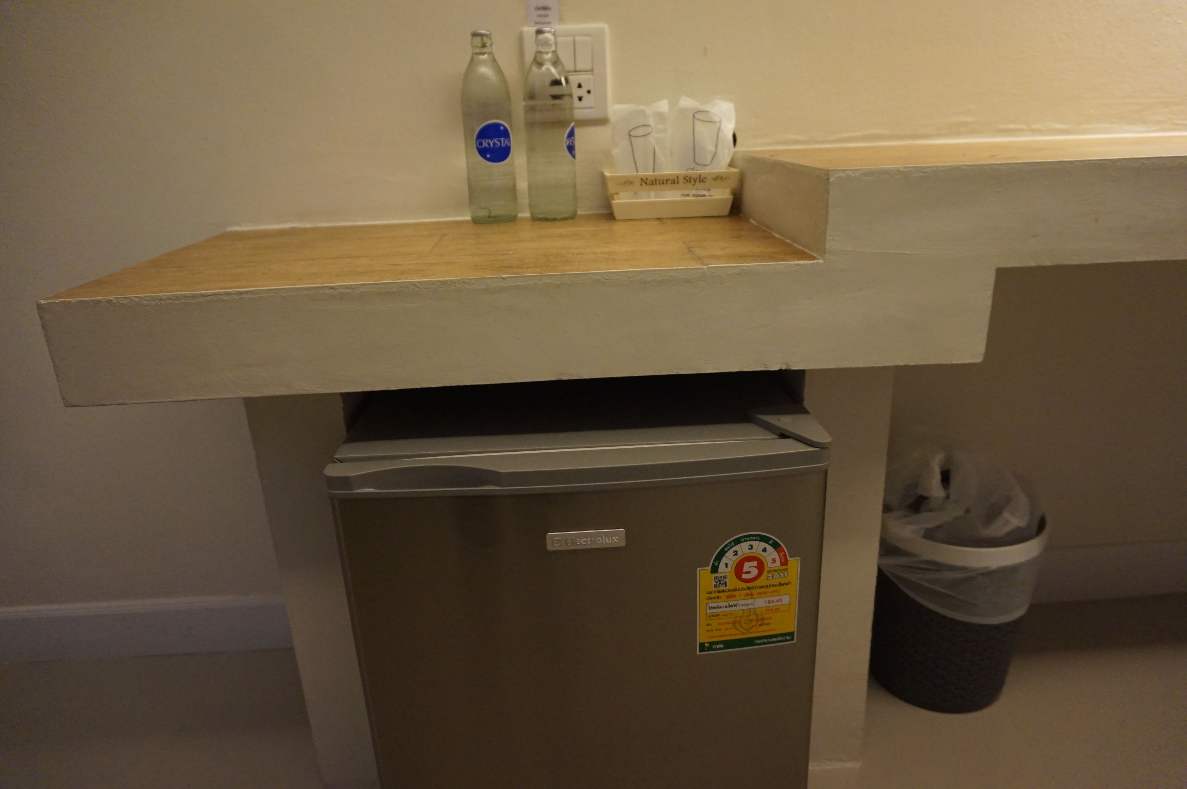 A compact refrigerator with a water dispenser, a trash bin, and a small shelf with bottles and a tissue box.