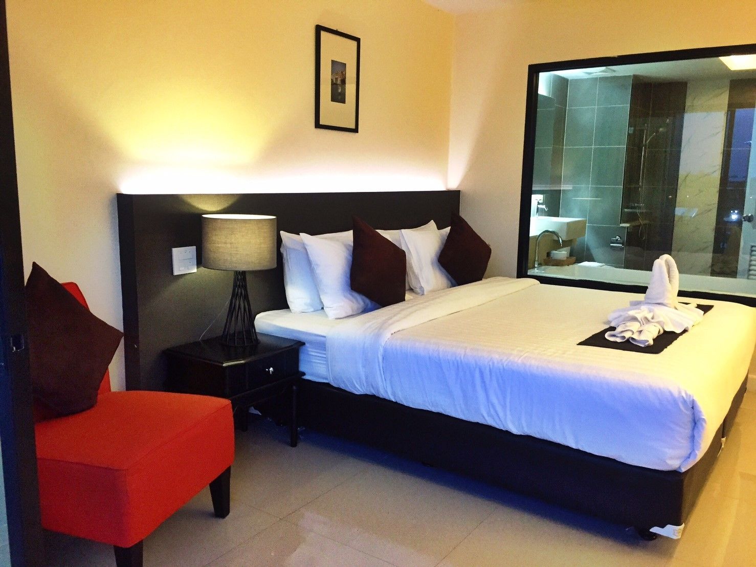 Une chambre d’hôtel avec un grand lit, une table de chevet avec une lampe, un fauteuil rouge et un grand miroir qui reflète la salle de bain.