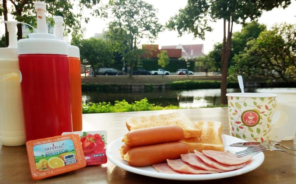 A breakfast setting with hot dogs, ham, toast, and condiments on a table with a view of a water body and trees.