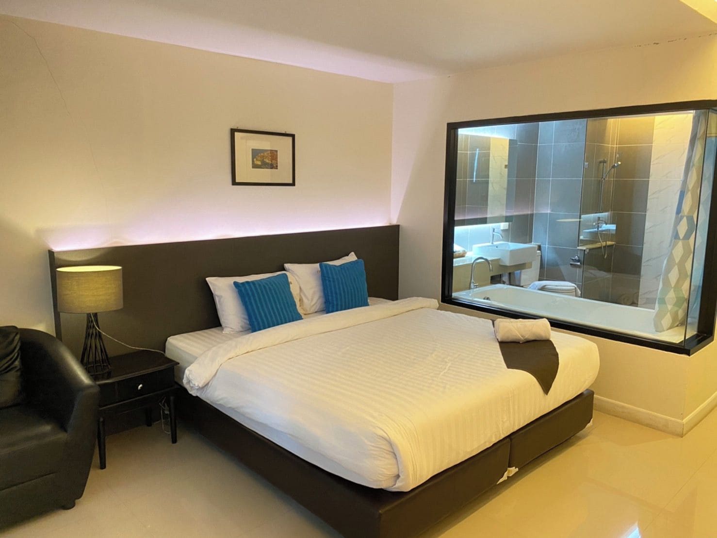 A modern hotel room with a large bed, a nightstand, a lamp, a black leather chair, and a view of the bathroom through a large window.