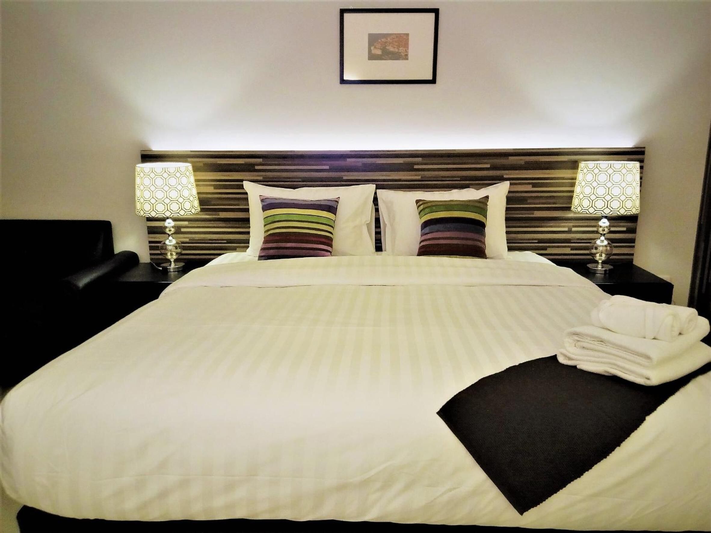 A neatly made bed with striped pillows, a headboard with horizontal wooden slats, two bedside lamps with patterned lampshades, and a framed picture on the wall.
