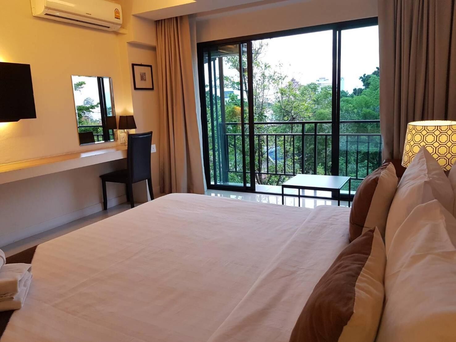 Habitación de hotel con cama, escritorio, silla, televisor y balcón con vistas a árboles y edificios.