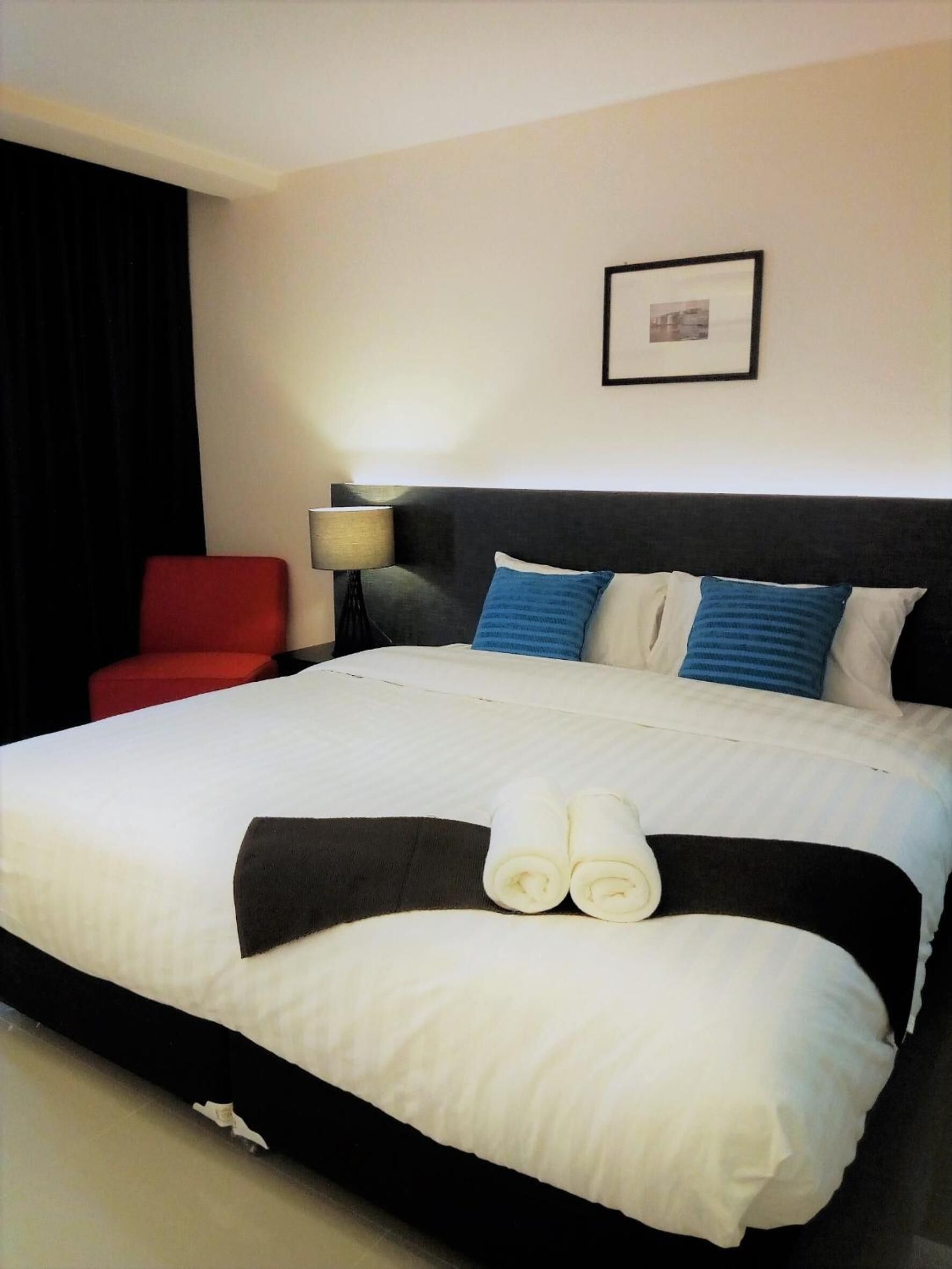 A hotel room with a large bed, a red chair, a nightstand with a lamp, and a framed picture on the wall.