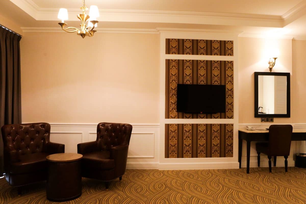 A hotel room with a large flat-screen TV mounted on a patterned wall, two leather armchairs, a small round table, a desk with a chair, and a chandelier.