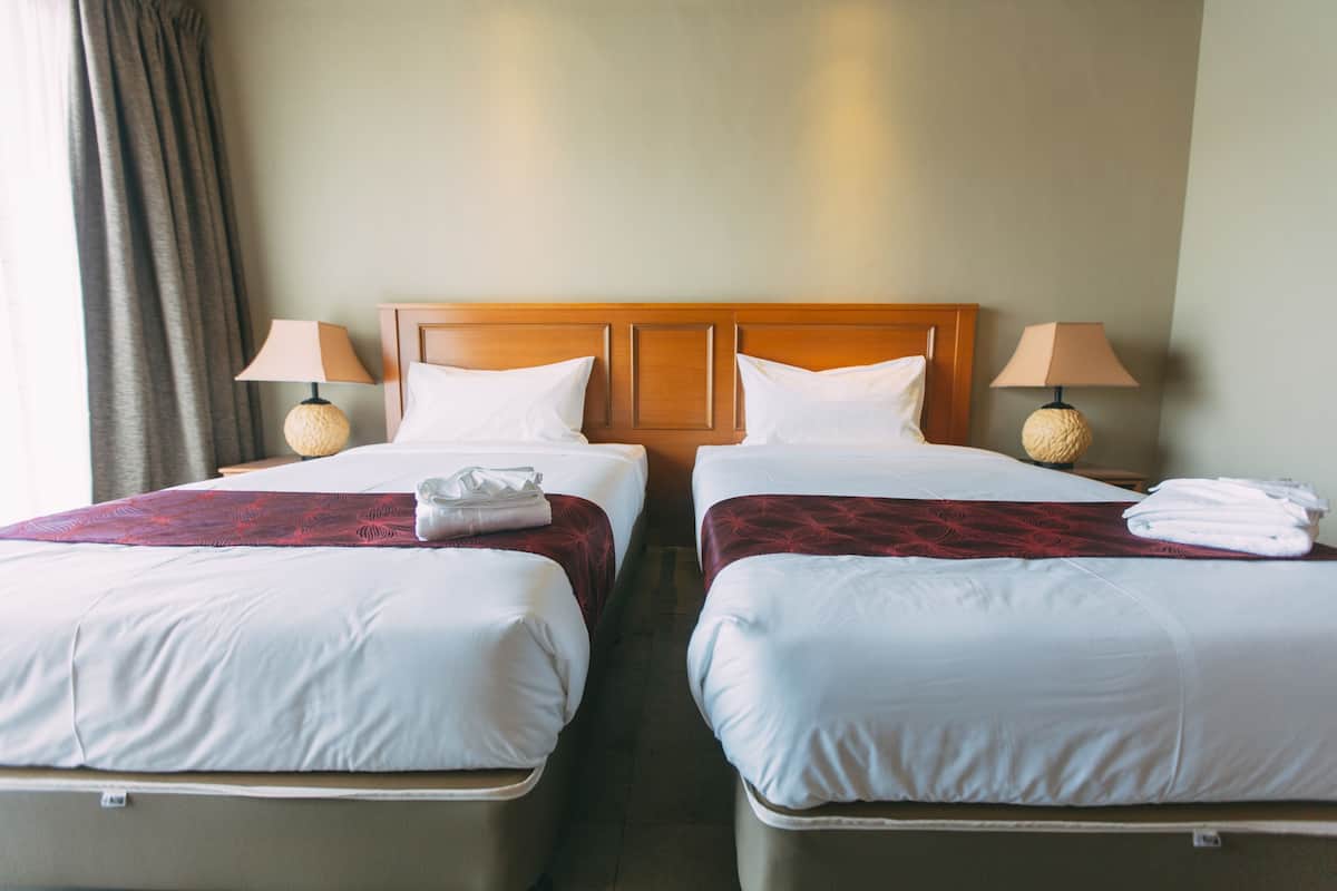 A hotel room with two beds, each with white linens and a wooden headboard. There are two bedside lamps with round bases and beige shades.