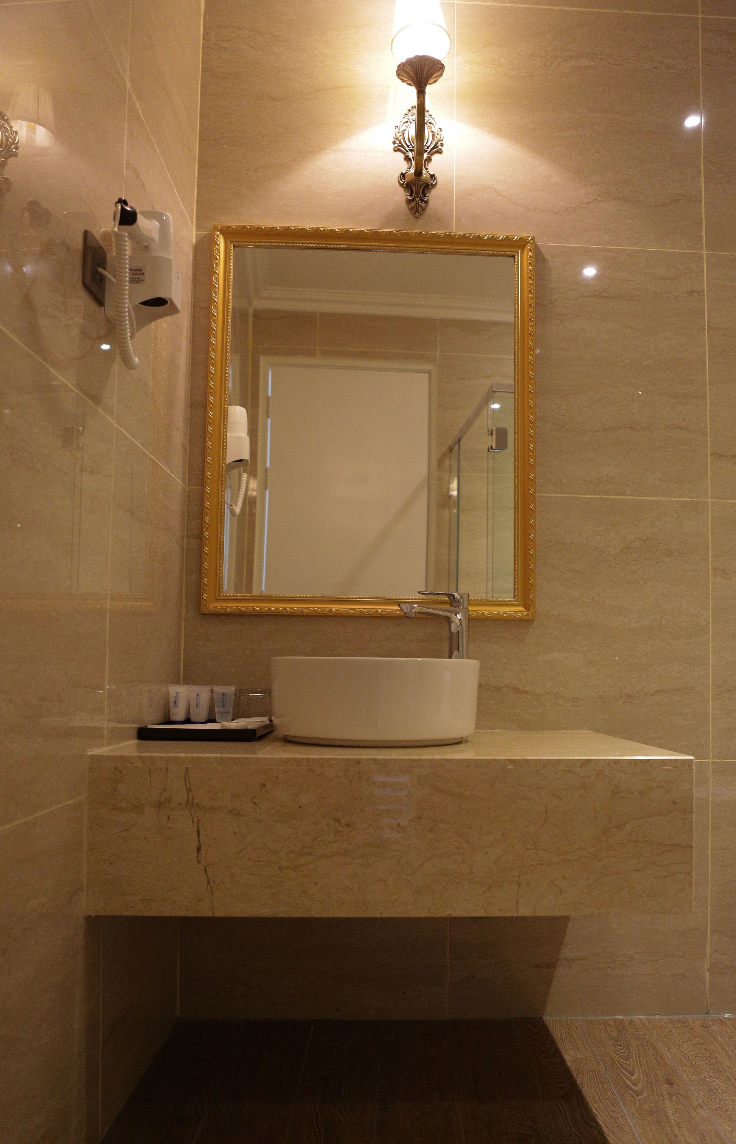 Un baño con un lavamanos blanco, un espejo con marco dorado, un aplique de pared y un secador de manos.
