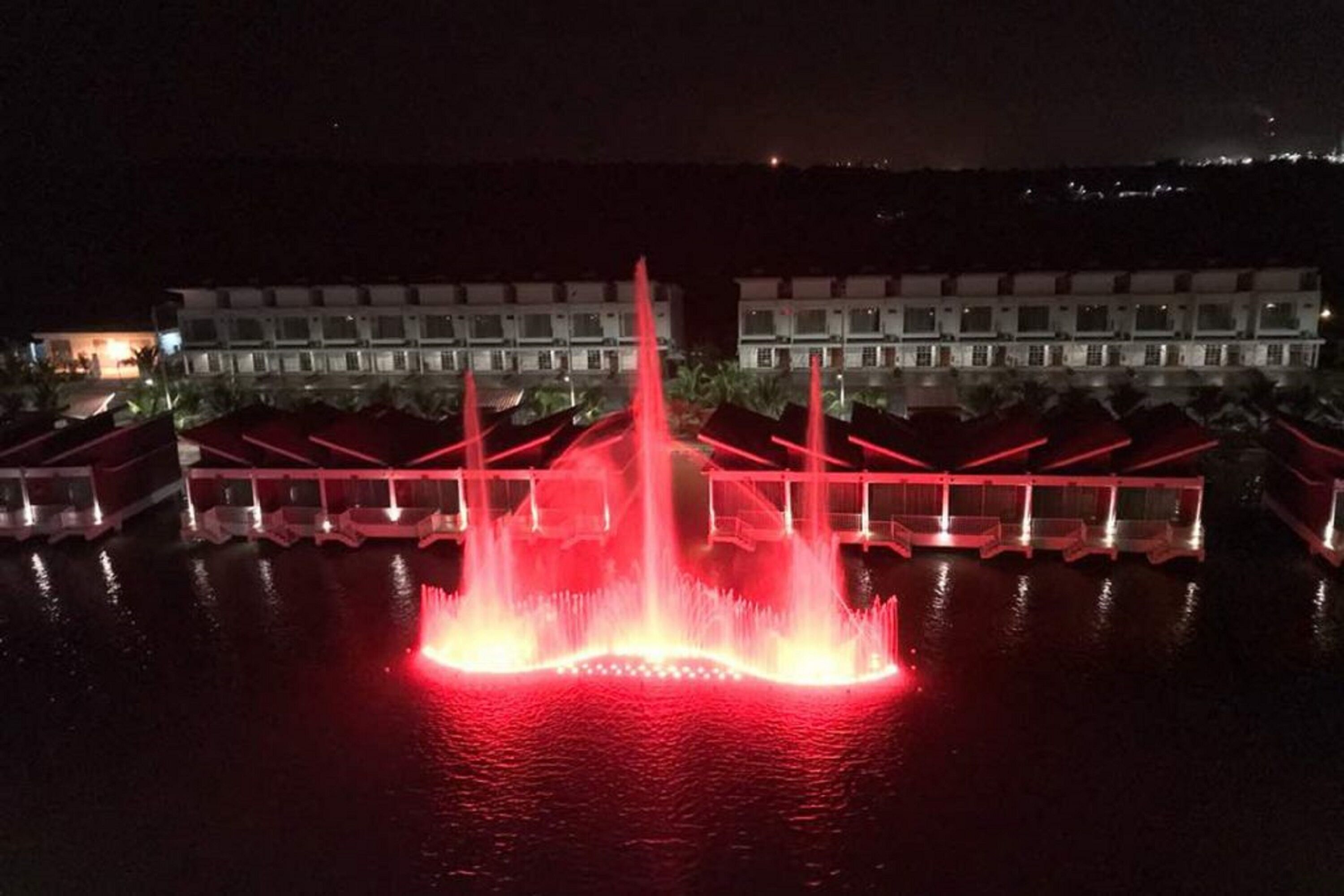 A nighttime view of a hotel with a central water fountain and a row of boats in the foreground.