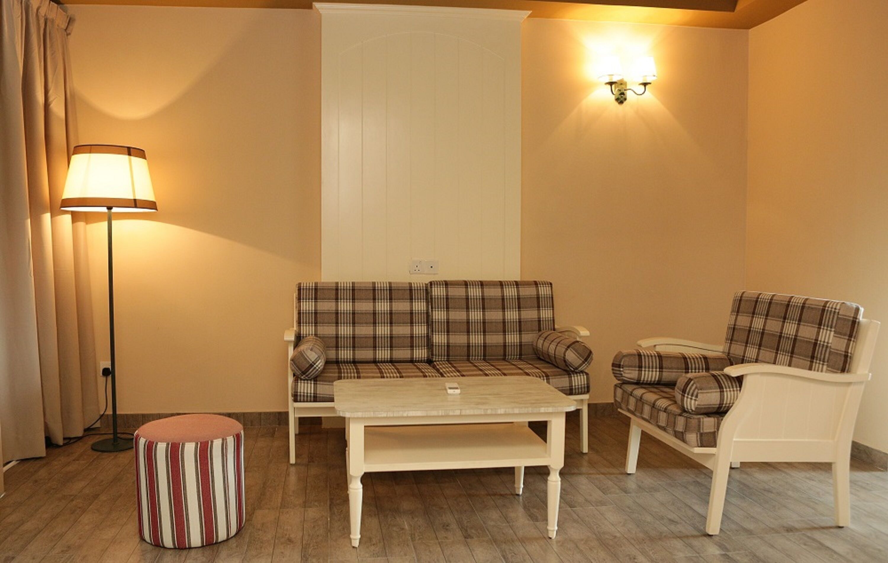 Una sala de estar acogedora con un sofá a cuadros, un sillón blanco, un reposapiés a rayas, una mesa de centro, una lámpara de pie y un aplique de pared.