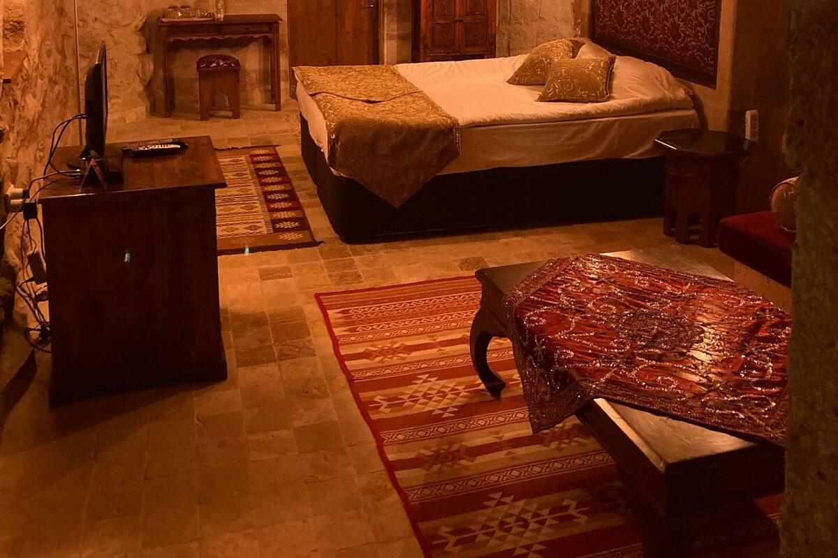 A cozy room with a stone arch ceiling, a bed with a headboard, a wooden dresser, a desk with a lamp, and a patterned rug.