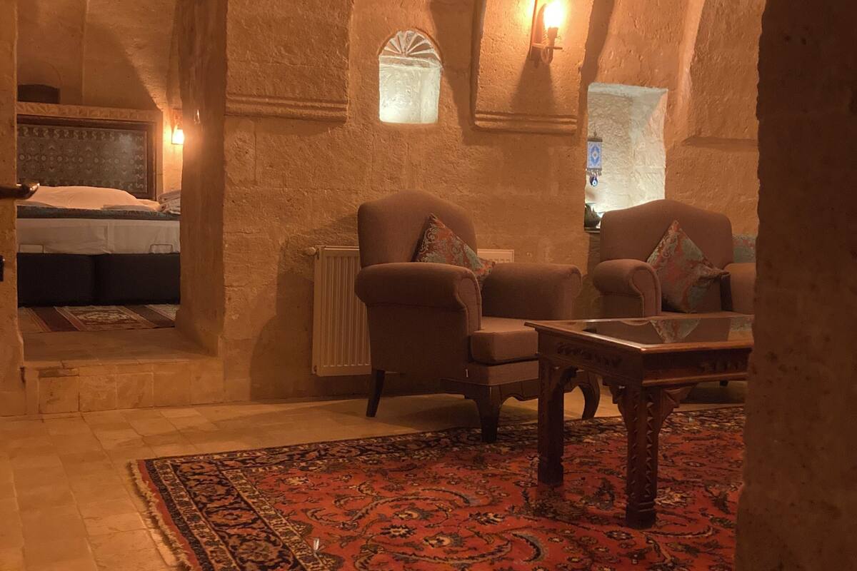 A dimly lit room with a high arched ceiling, a bed with patterned headboard, a sofa with a cushion, a small table, and a patterned rug on the floor.