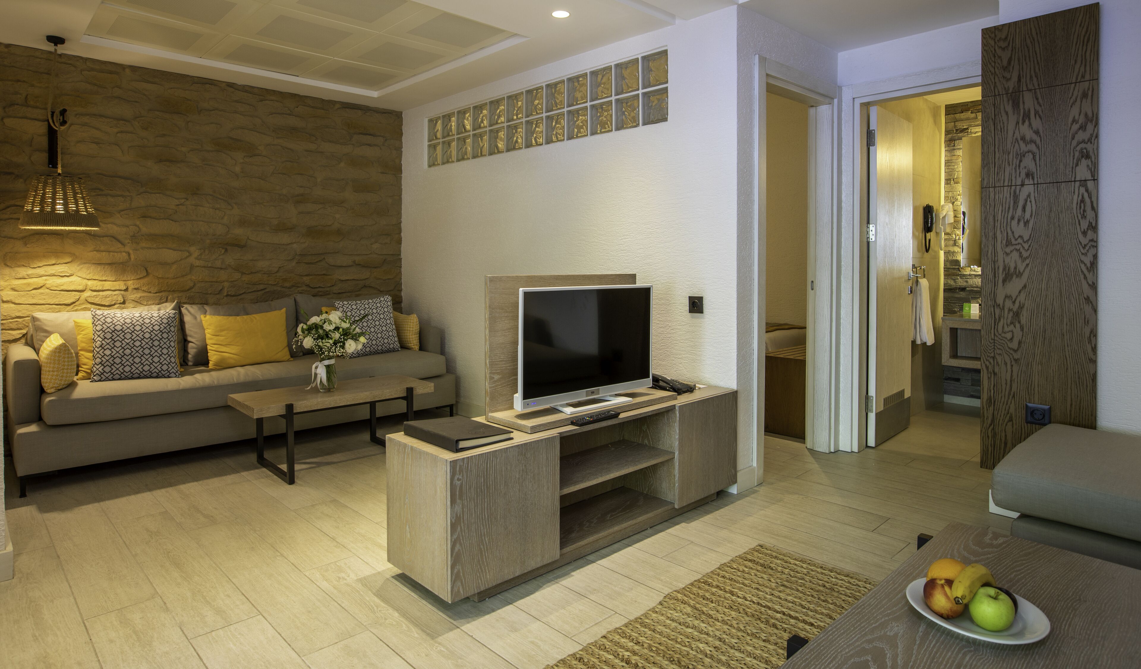 A modern living room with a flat-screen TV on a wooden stand, a sofa with patterned cushions, a stone wall, and a wooden door.