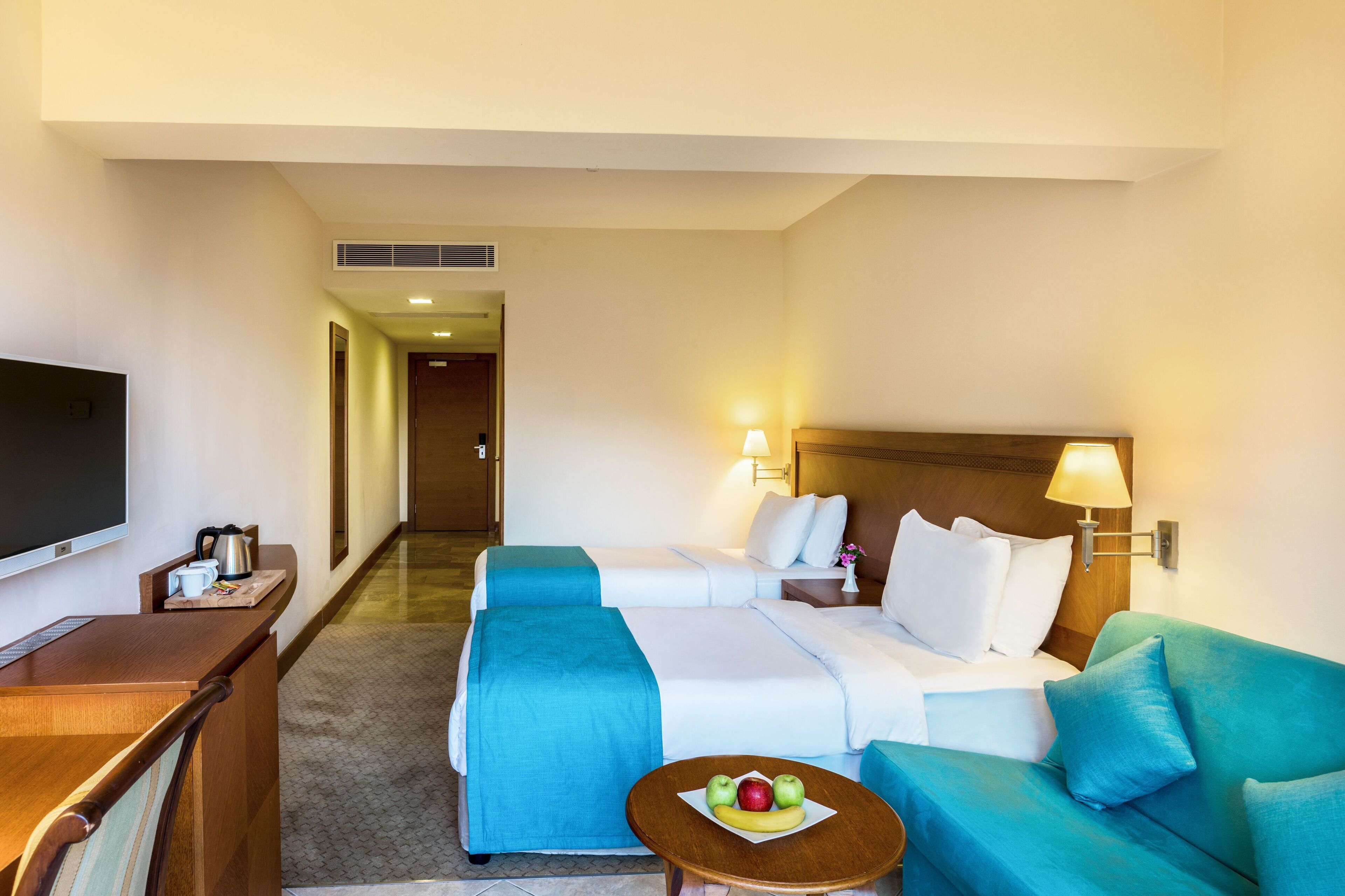 Une chambre d’hôtel avec un lit, un canapé, une télévision, une table basse avec des fruits et un bureau avec une lampe.