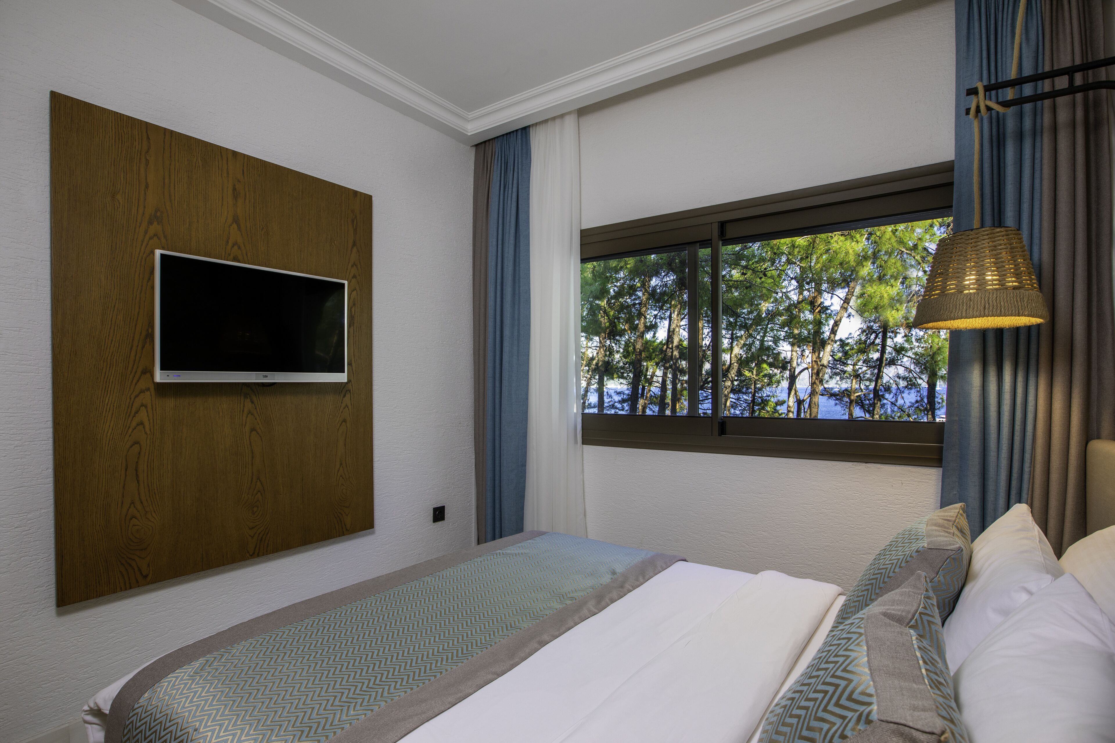 Ein Schlafzimmer mit Bett, montiertem Fernseher, Fenster mit Blick auf Bäume und einer Hängelampe.