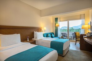 A hotel room with two beds, a desk with a lamp, a chair, and a view of the outdoors.