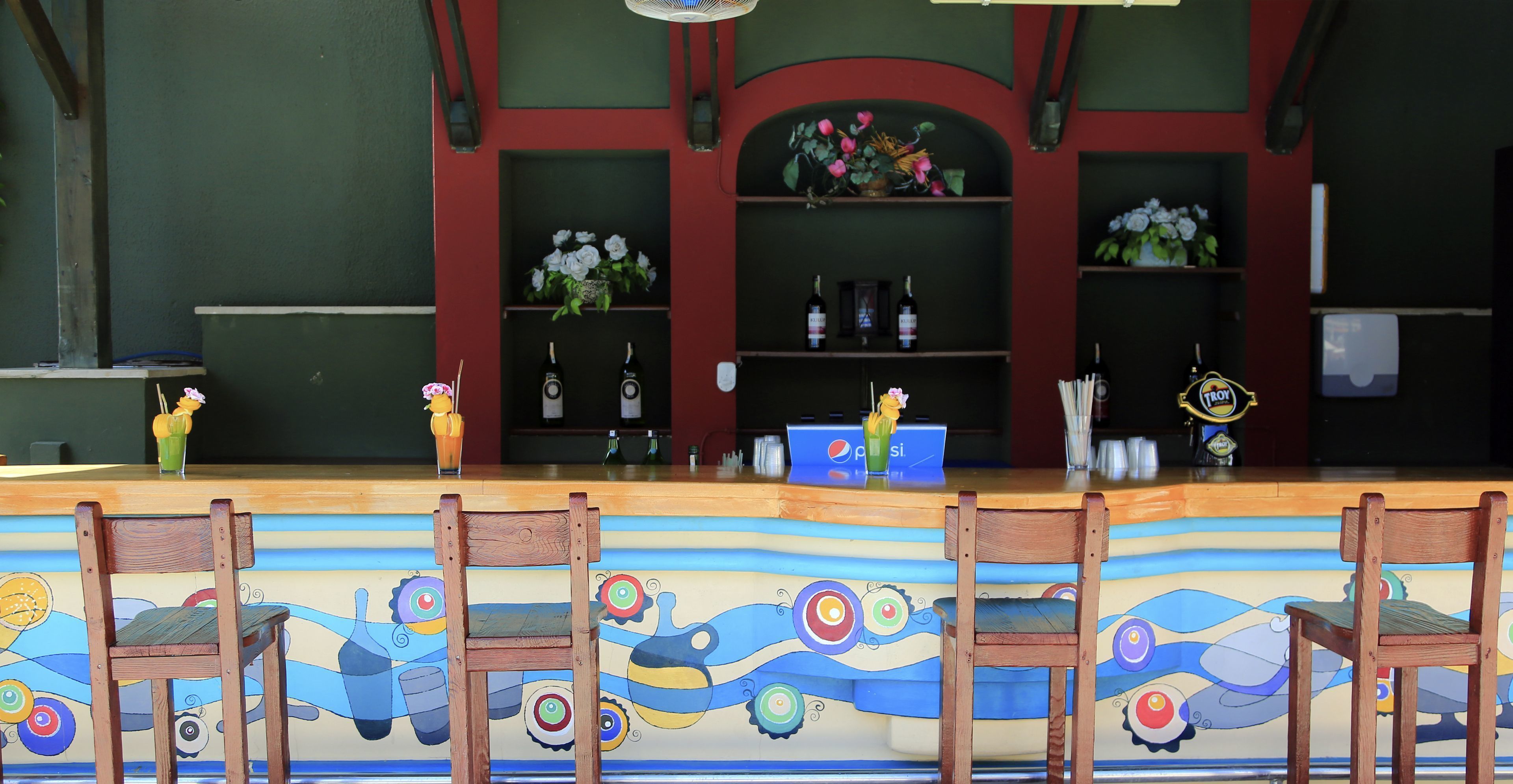 A bar area with wooden chairs, a colorful mural, shelves with flowers and bottles, and a Pepsi sign.