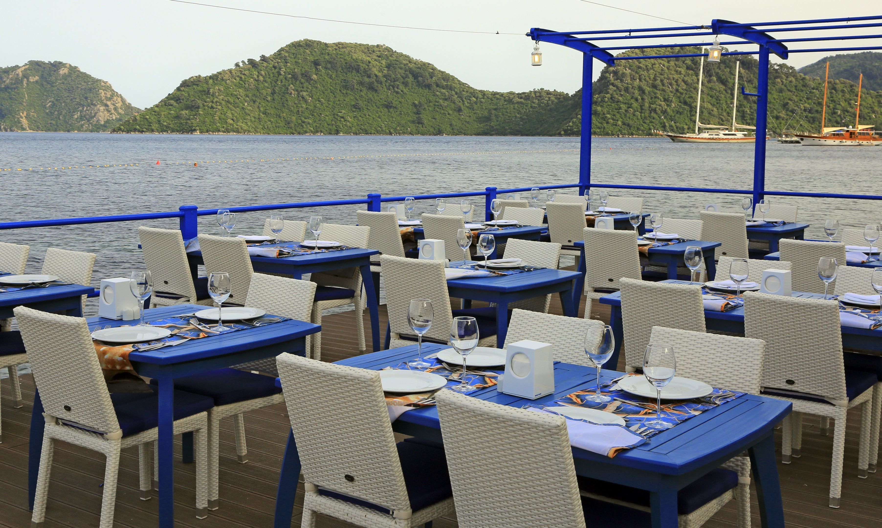 A restaurant deck with tables and chairs, overlooking a body of water with boats and hills in the background.