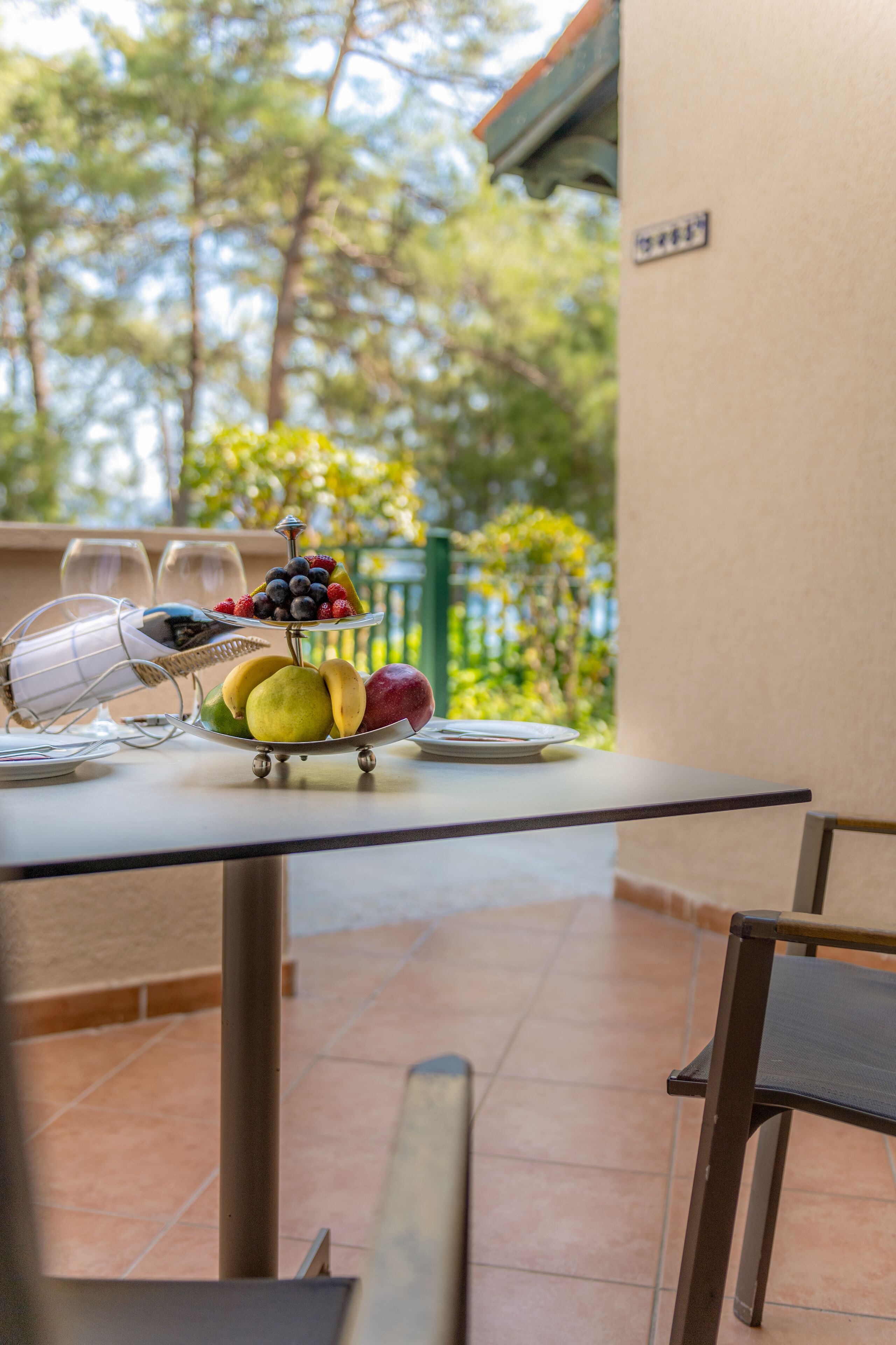 A table with a fruit platter, wine glasses, and a chair on a balcony with a tiled floor and a green railing.