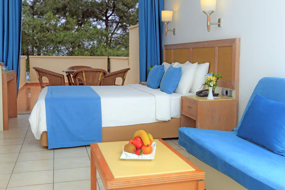 Quarto de hotel com cama, sofá, mesinha com frutas, criado-mudo, cadeira e vista para árvores.