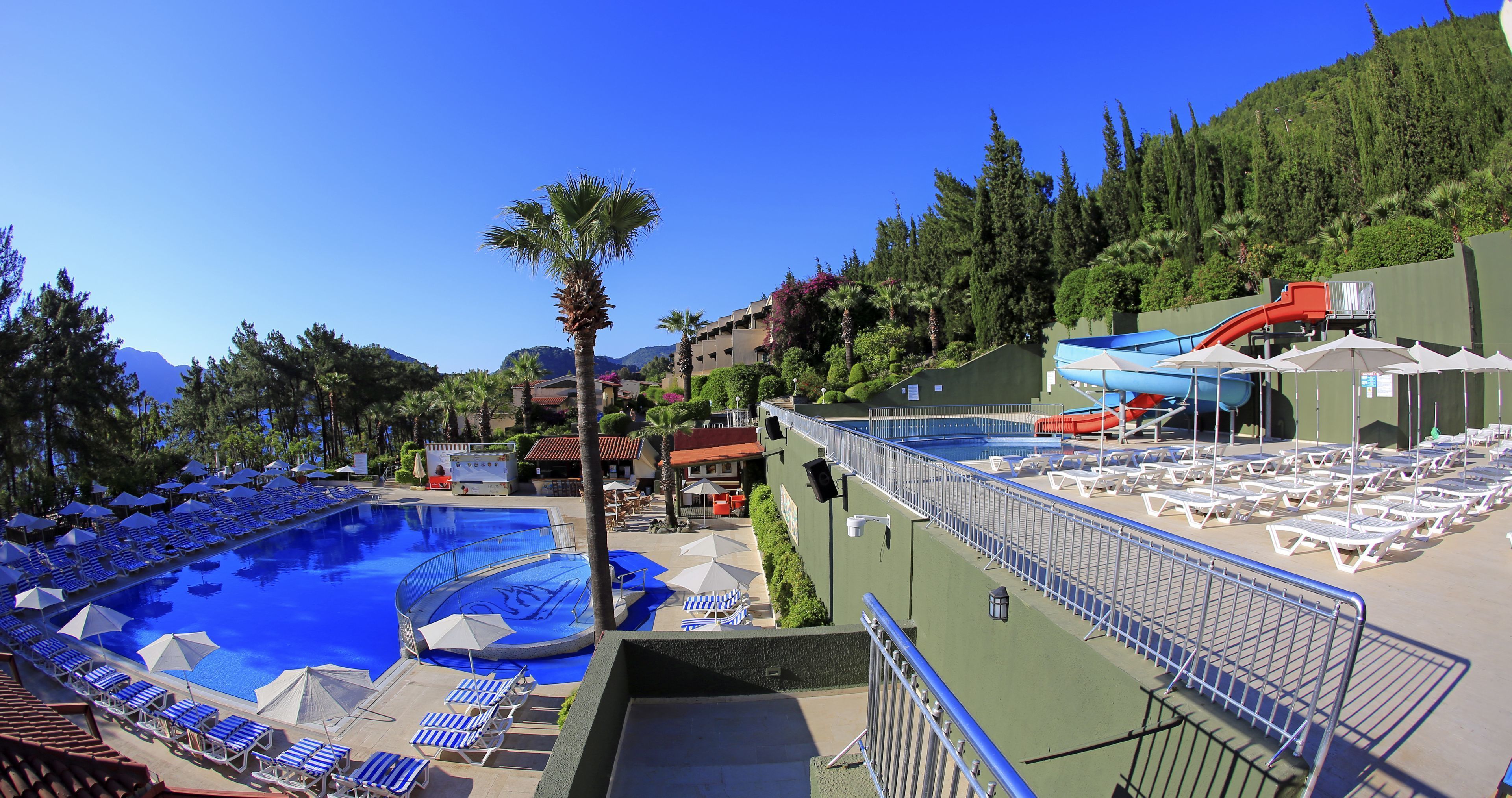 Un espace piscine aménagé dans un complexe hôtelier, avec des chaises longues et des parasols, un toboggan aquatique et une vue sur les montagnes.
