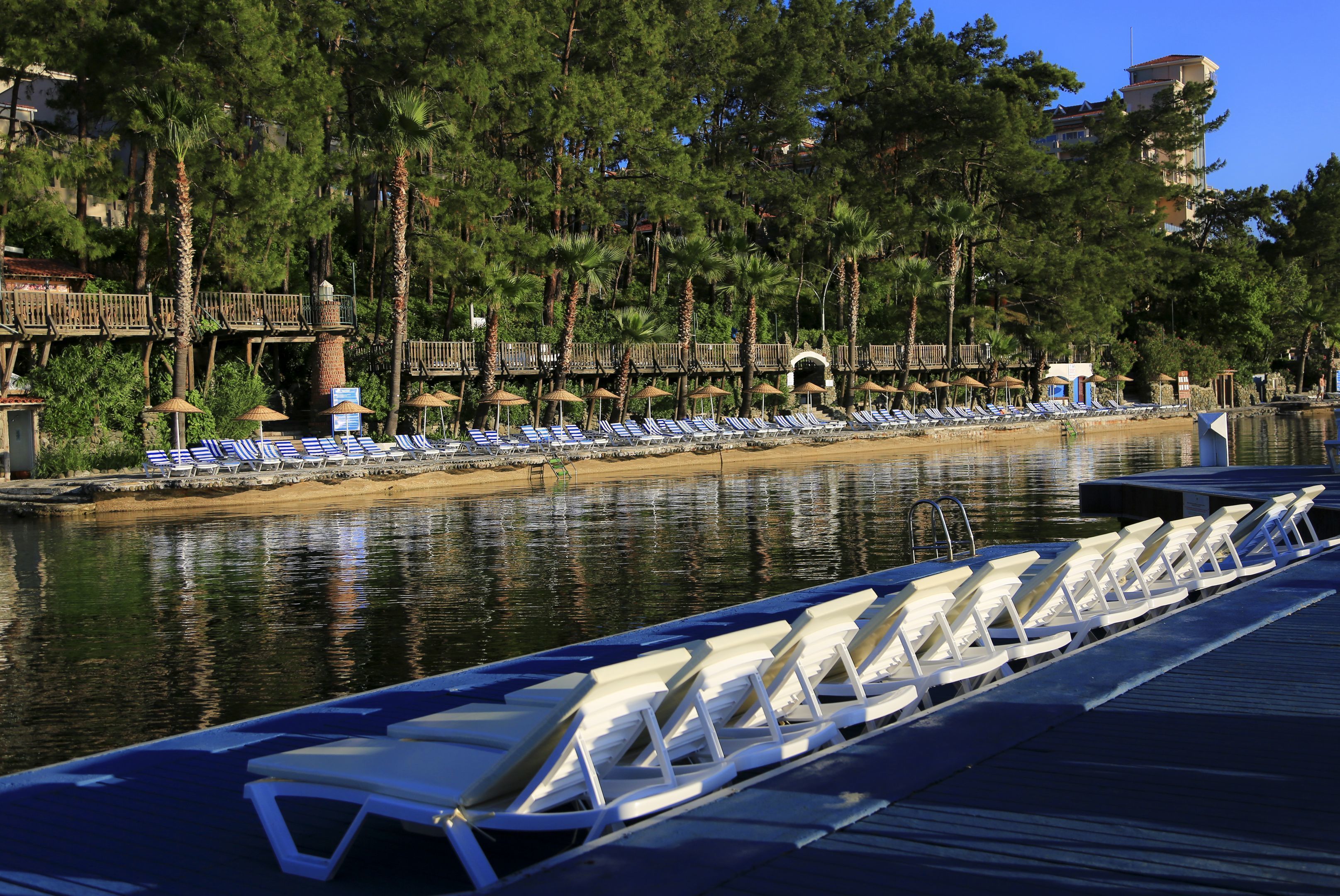 A waterfront area with lounge chairs and umbrellas along the shore, a wooden bridge, and a building in the background.