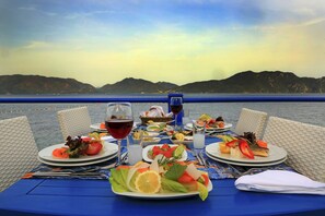 A table set for a meal with plates of food, glasses of wine, and a view of the sea and mountains in the background.