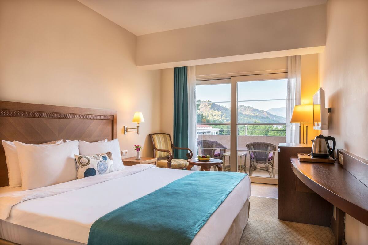 Quarto de hotel com uma cama grande, uma escrivaninha, uma cadeira, varanda com vista para as montanhas e janela com cortinas.